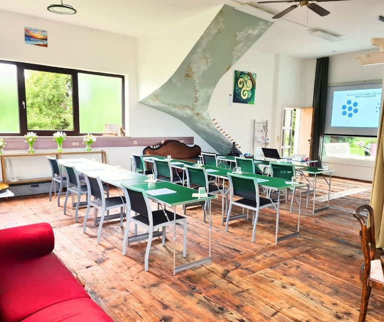 Heller Schulungsraum mit mehreren grünen und weißen Tischen, bestückt mit Stühlen, auf altem Holzboden. Im Hintergrund Fenster mit Blick auf grünes Laub, eine Projektionsfläche und dekorative Gemälde an der Wand.